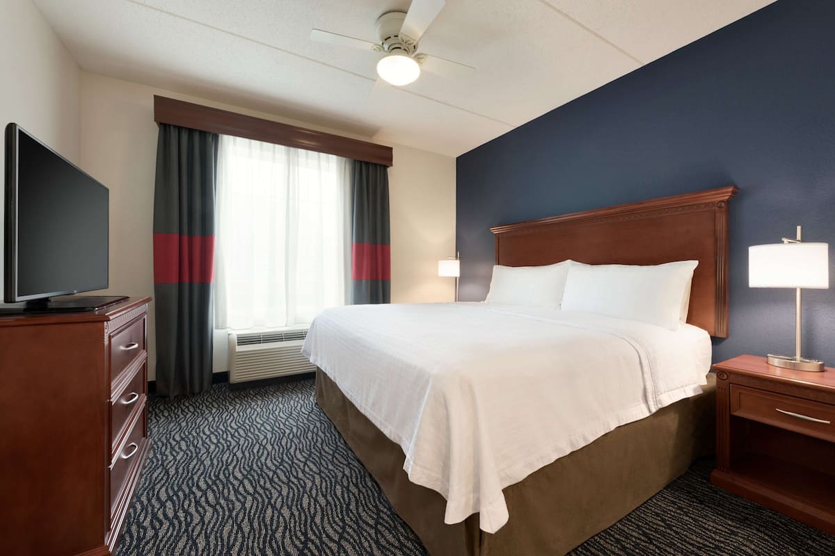 A hotel room with a large bed, a flat-screen TV, a desk with a lamp, and a window with curtains.