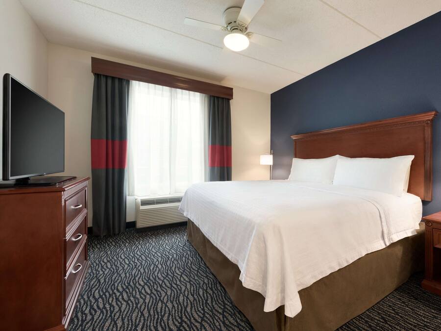 A hotel room with a large bed, a flat-screen TV, a desk with a lamp, and a window with curtains.