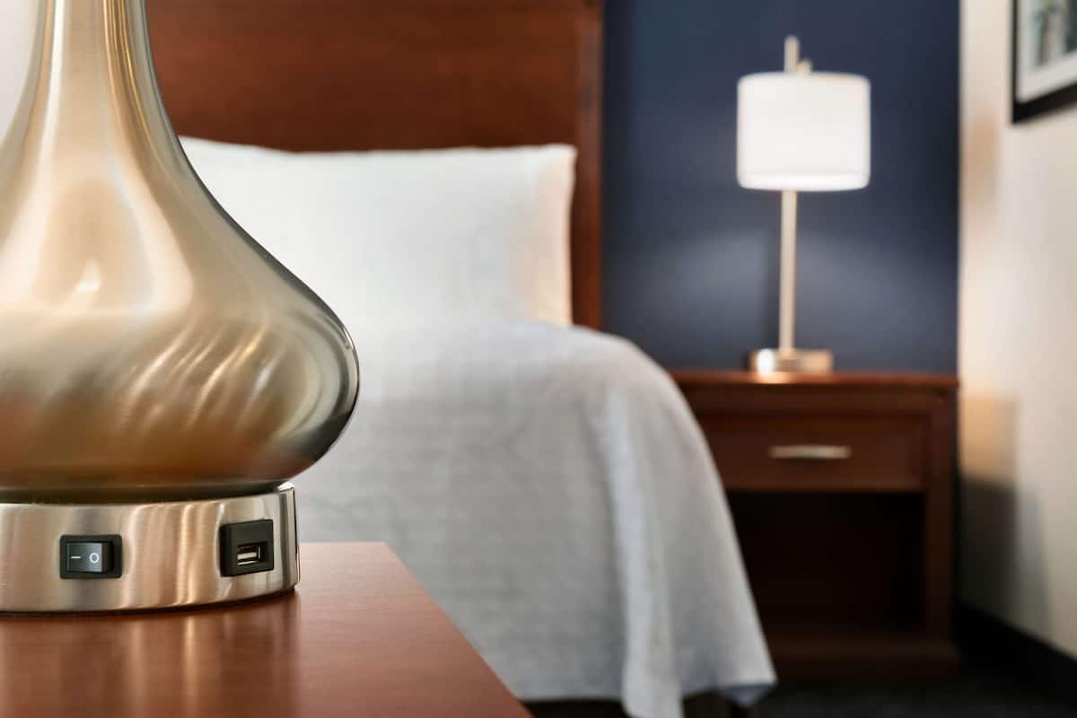 A modern hotel room with a bedside table featuring a decorative lamp, a neatly made bed, a nightstand with a lamp, and a wooden headboard.