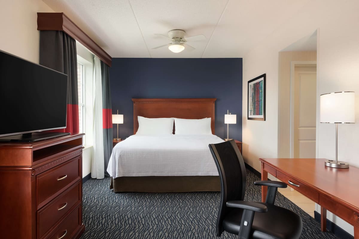 A hotel room with a large bed, a TV on a wooden dresser, a desk with a chair, and a ceiling fan.