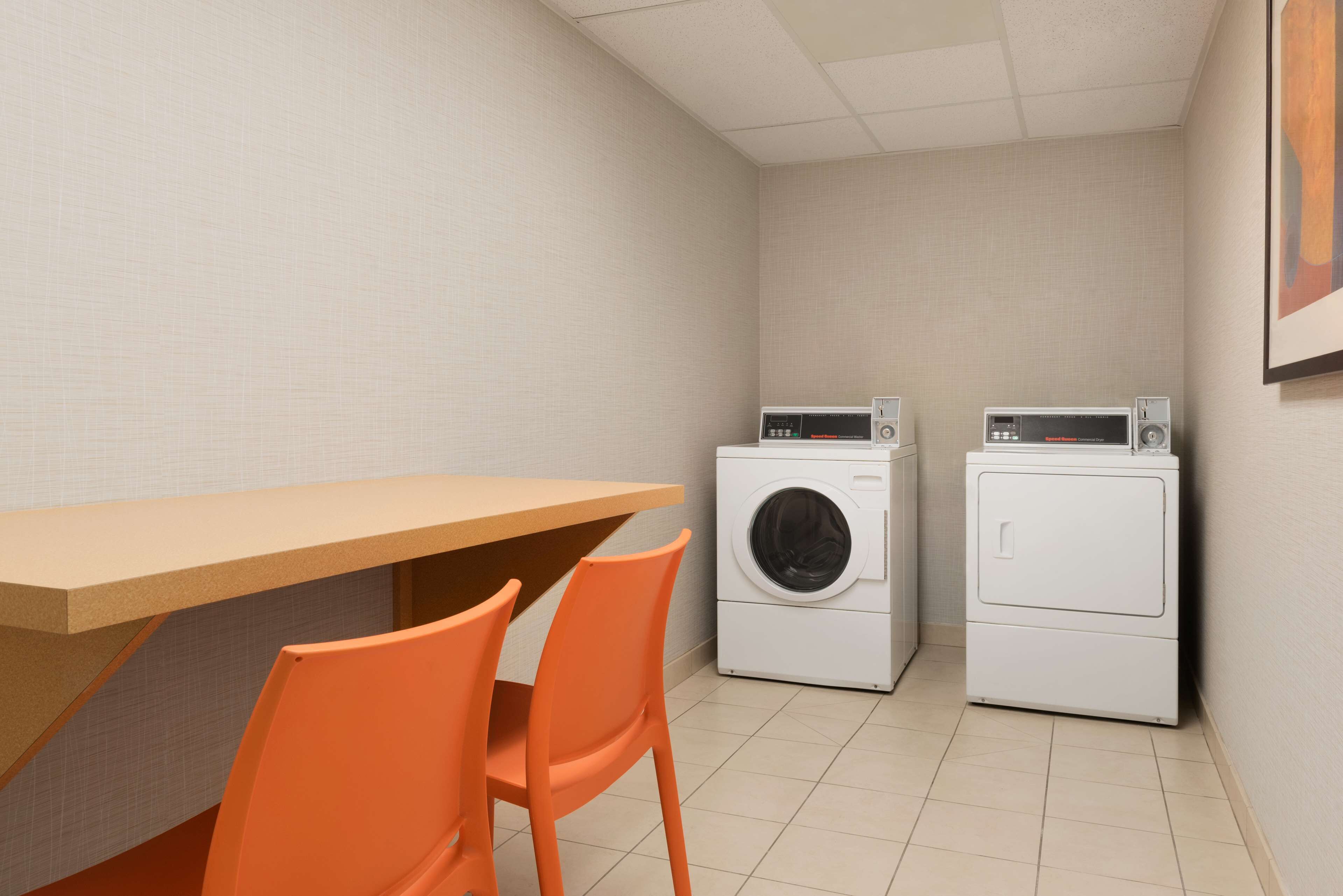 A laundry room with a wooden table and two orange chairs, two washing machines, and a tiled floor.