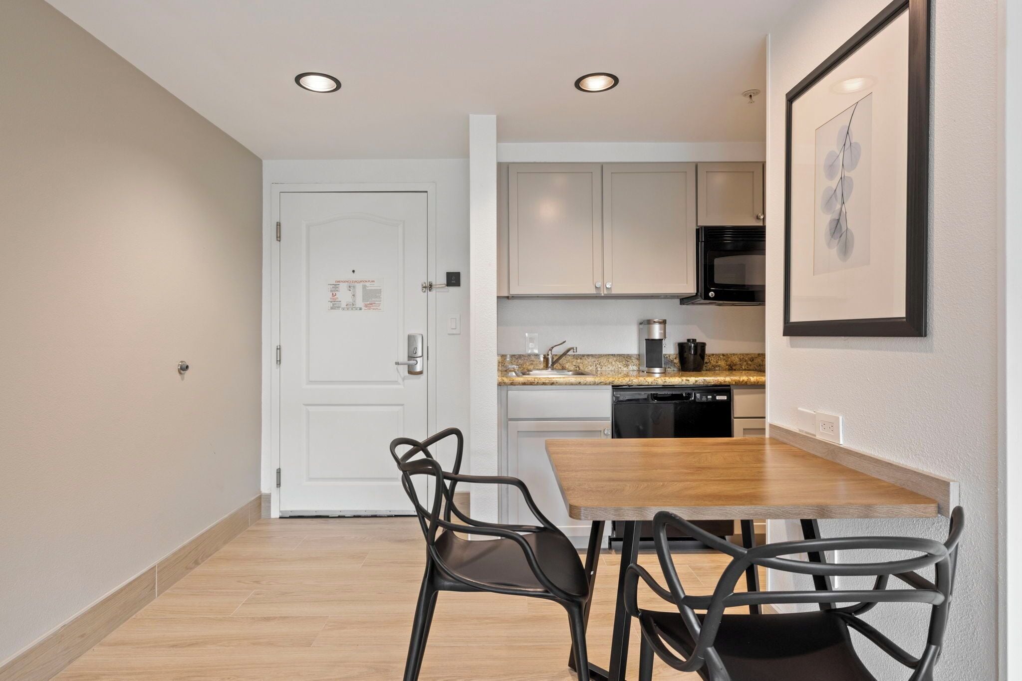 A compact kitchen with a dining area, featuring a white door, wooden table, and black chairs.