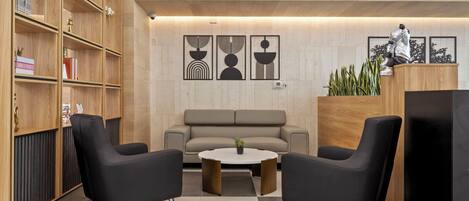 Un vestíbulo moderno con un sofá, dos sillones negros, una mesa de centro, estanterías de madera con libros y objetos decorativos, y tres obras de arte enmarcadas en la pared.