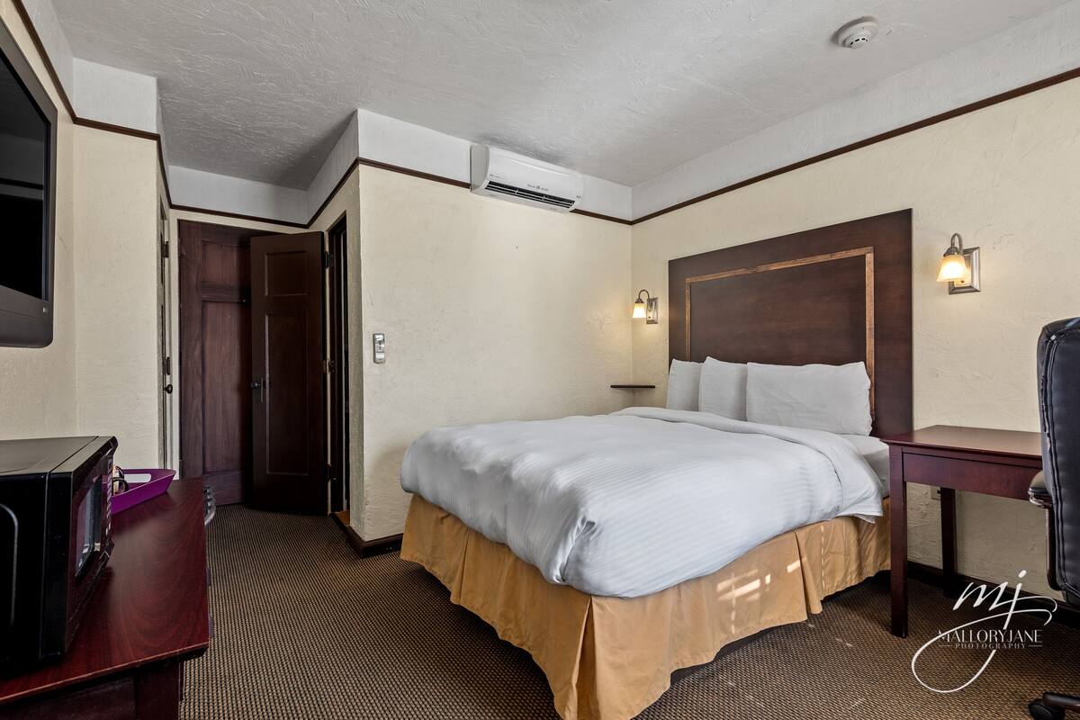 A hotel room with a bed, a desk, a chair, a nightstand, a lamp, and an air conditioning unit.