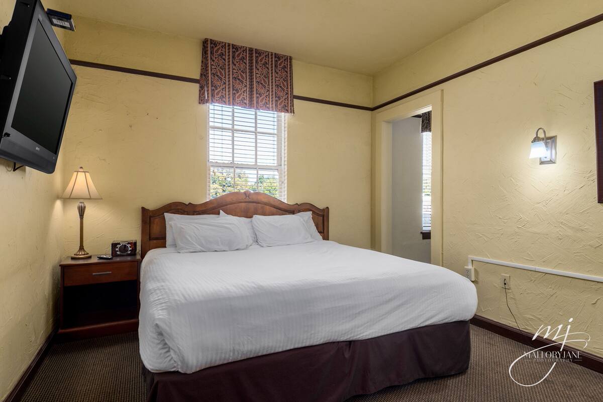 A hotel room with a large bed, a bedside table with a lamp, a flat-screen TV mounted on the wall, and a window with blinds.
