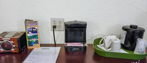 Un escritorio con un reloj digital, una cafetera, una bandeja verde con tazas y una caja para aventuras todoterreno.