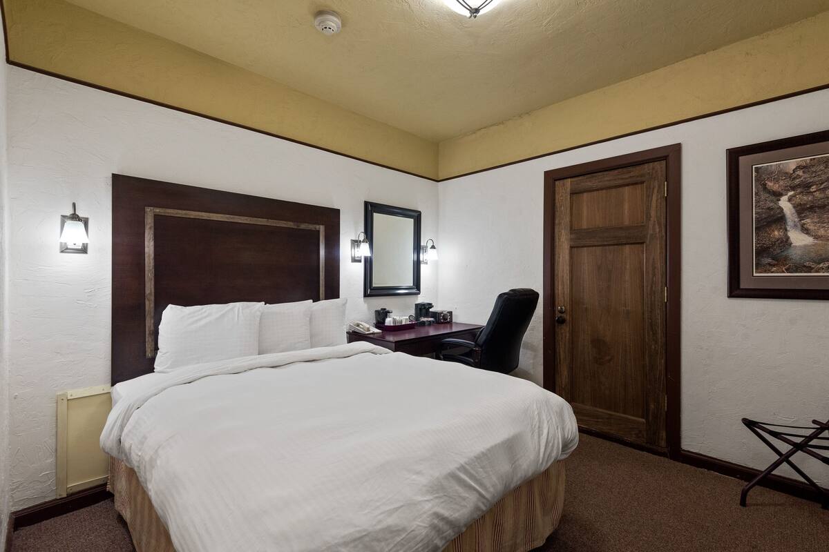 A hotel room with a large bed, a desk with a chair, a wooden door, and a framed picture on the wall.
