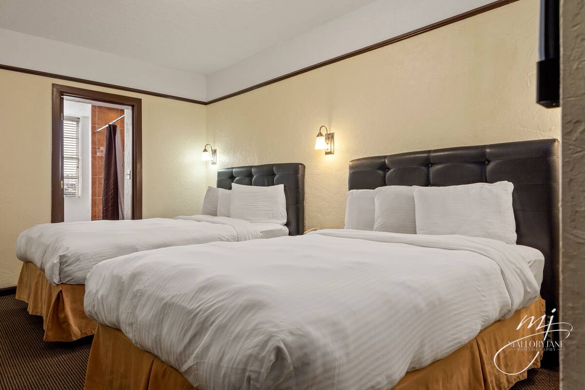 A hotel room with two beds, each with white linens and a dark headboard. There are wall-mounted light fixtures on either side of the beds.