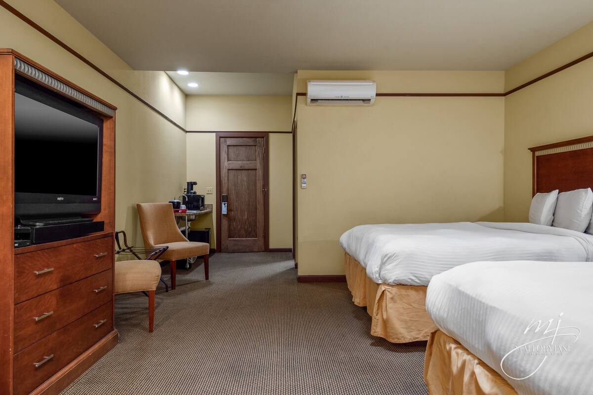 A hotel room with two beds, a TV, a desk, and an air conditioning unit.