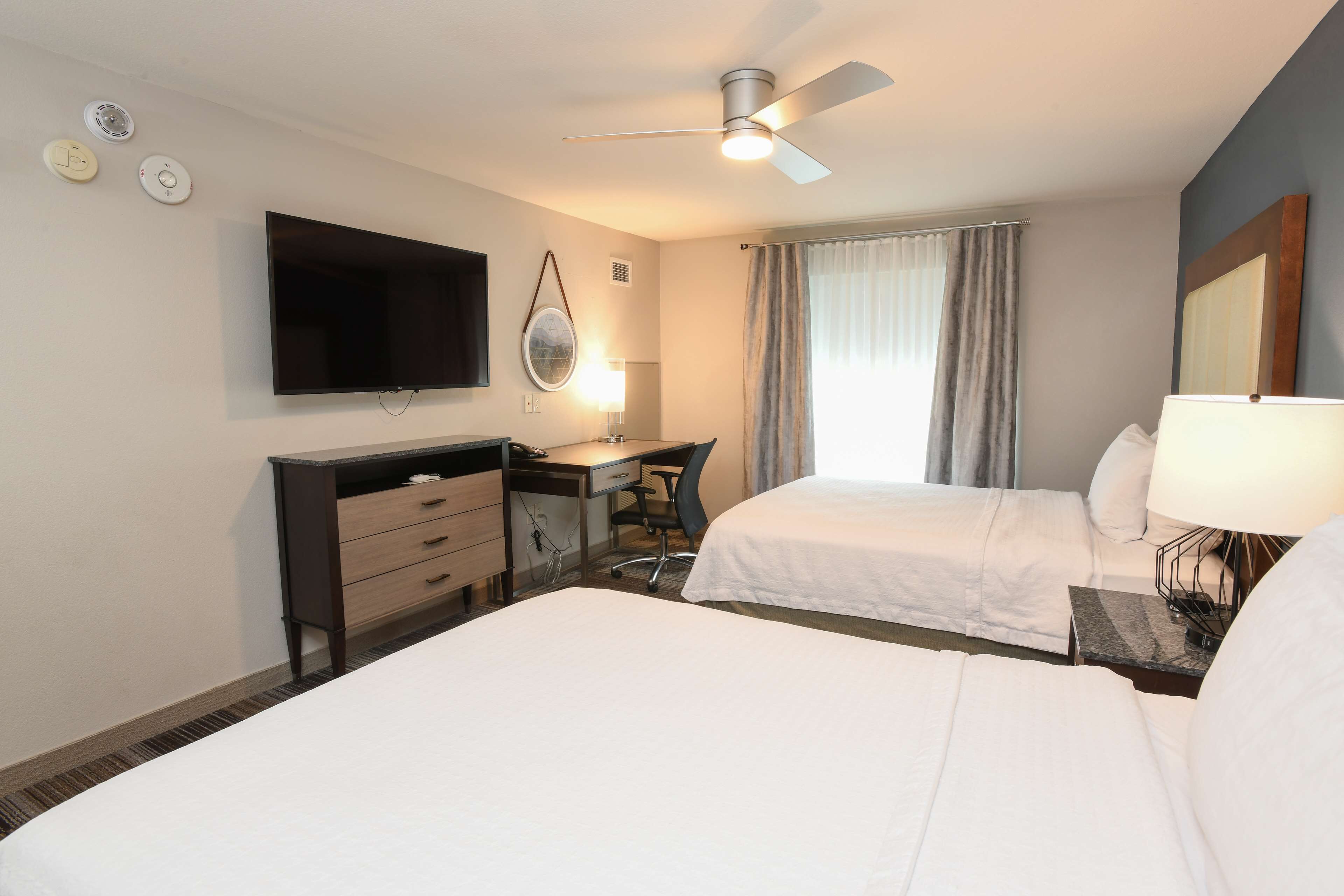 A hotel room with two beds, a desk, a chair, a television, a ceiling fan, and a window with curtains.