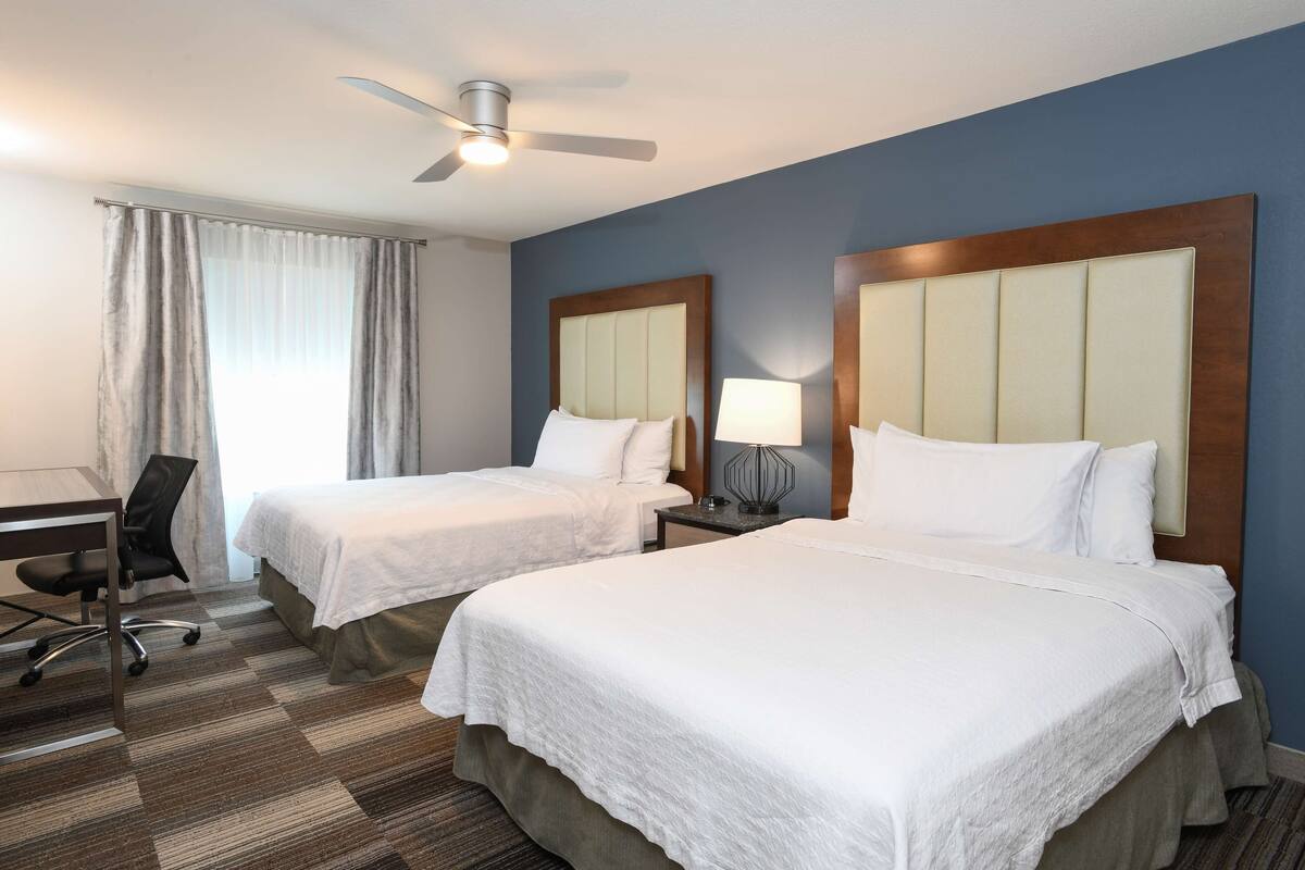 A hotel room with two beds, a desk, a chair, a ceiling fan, and a window with curtains.