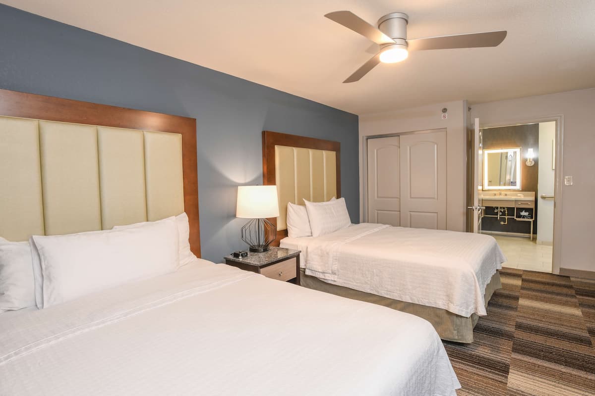 A hotel room with two beds, a ceiling fan, a nightstand with a lamp, and a bathroom visible through an open door.