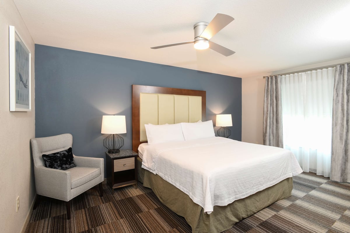 A hotel room with a bed, a chair, a nightstand, a lamp, a ceiling fan, and a window with curtains.