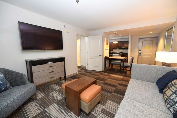 A modern living room with a flat-screen TV, a sofa, a coffee table, and a kitchen area in the background.