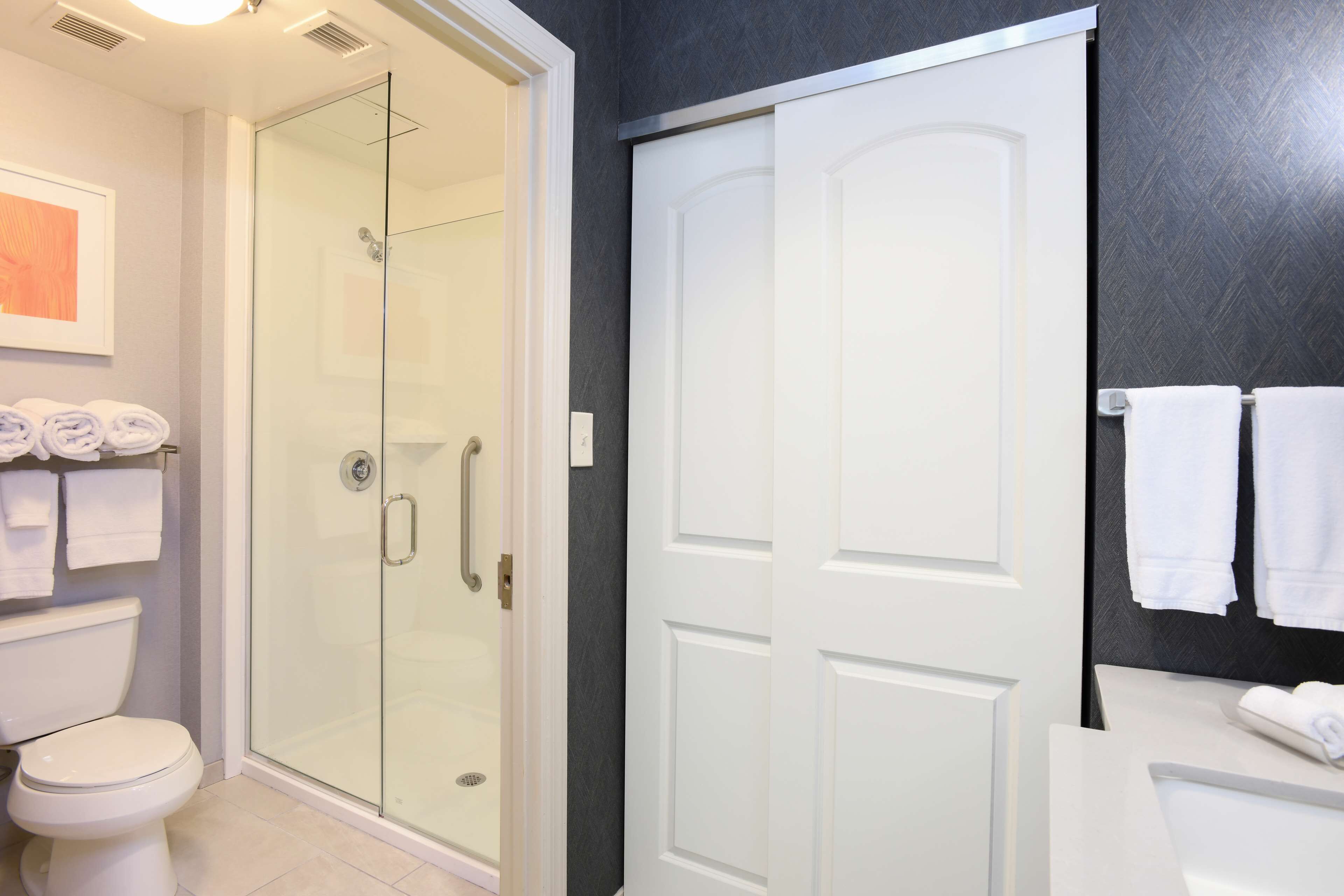 Un baño moderno con cabina de ducha de vidrio, inodoro y lavamanos blancos, y toallas.
