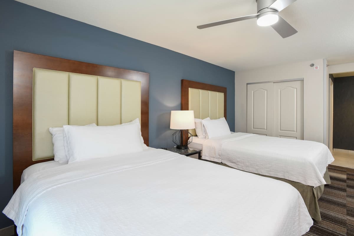 A hotel room with two beds, a ceiling fan, a nightstand with a lamp, and a door.