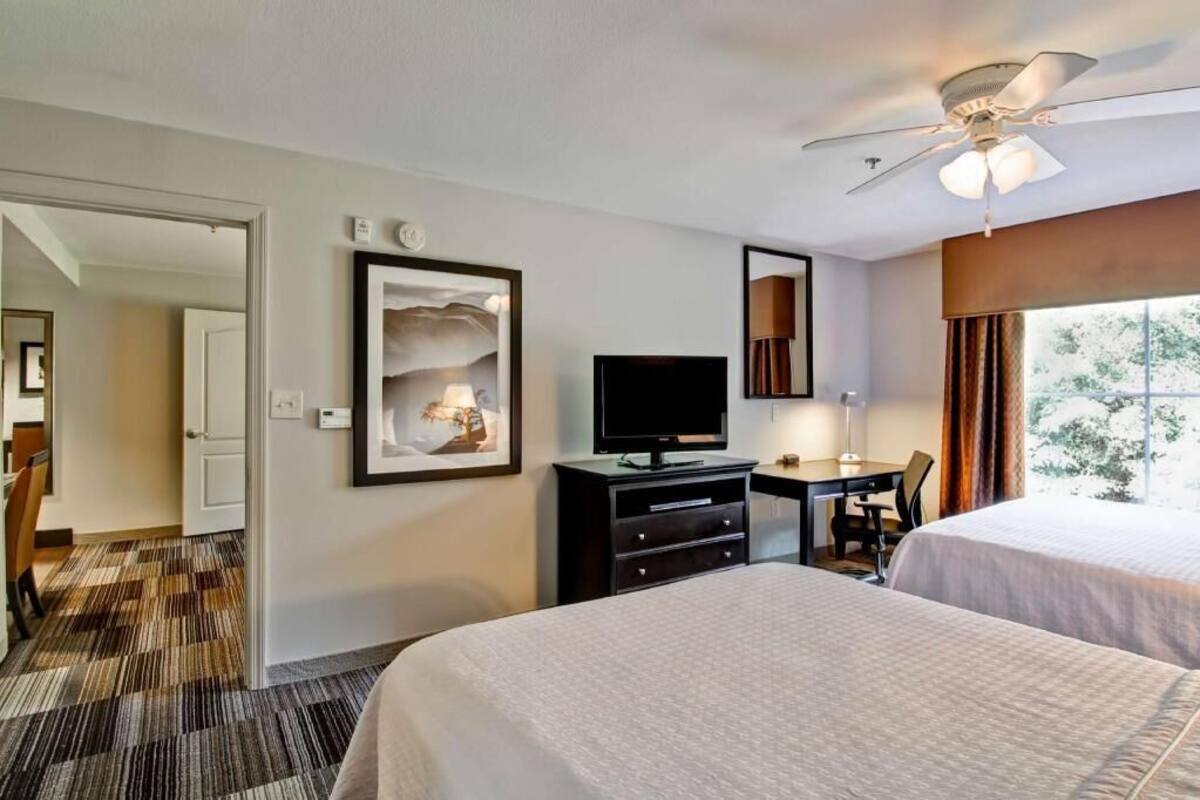 A hotel room with two beds, a desk with a chair, a television, a ceiling fan, and a large window.