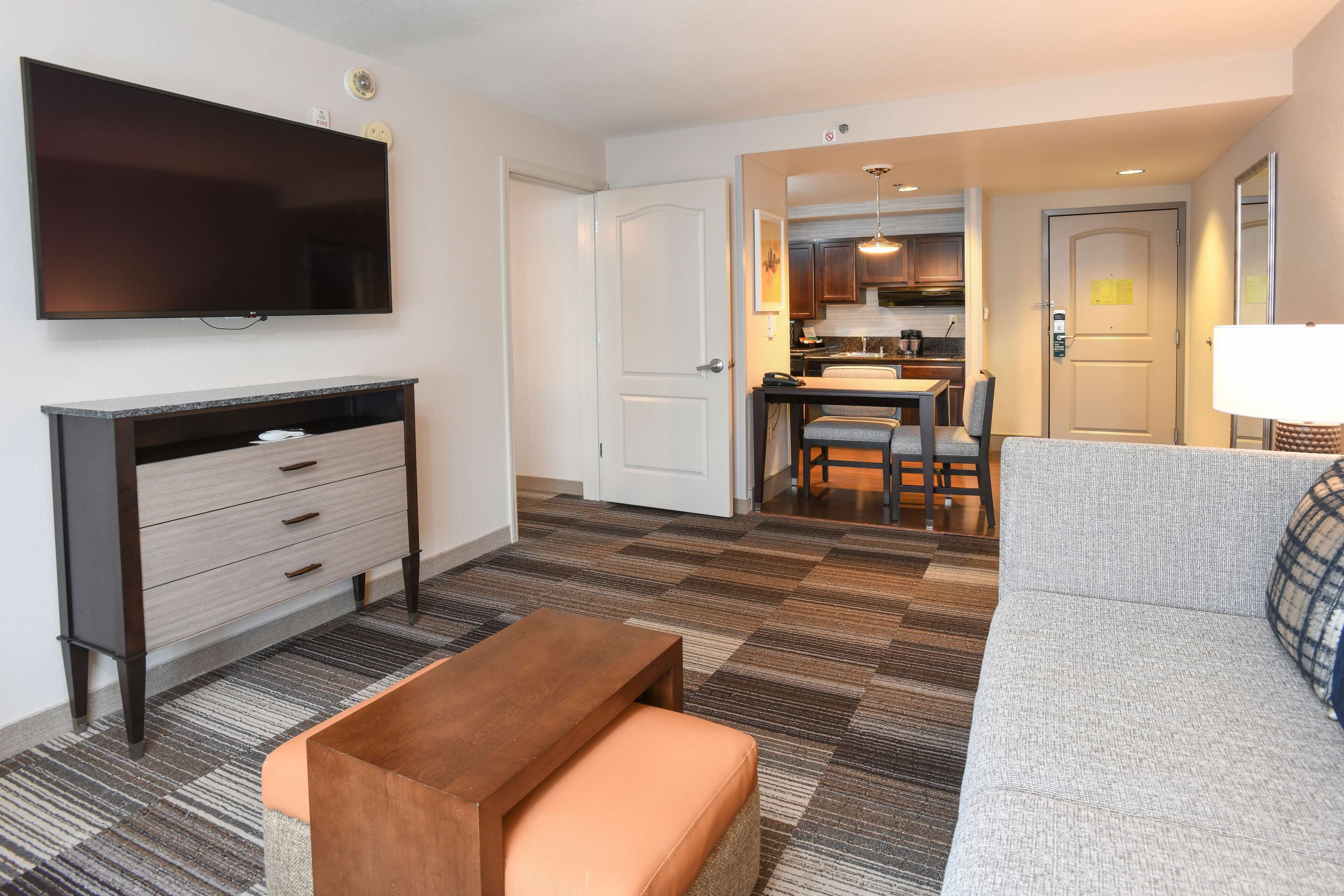 A modern hotel room with a flat-screen TV, a wooden bench, a grey sofa, and a kitchen area visible through an open door.