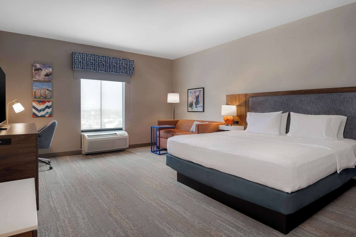 A hotel room with a large bed, a desk, a chair, a standing lamp, and artwork on the walls.