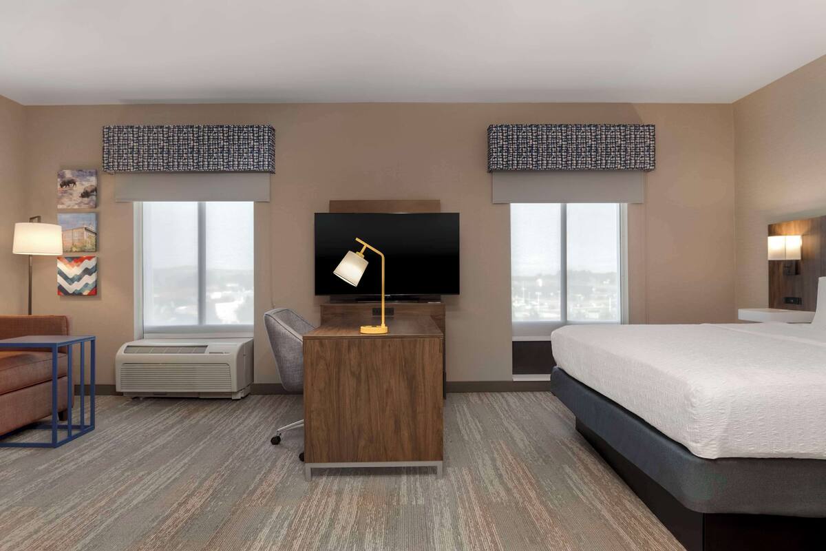 A hotel room with a bed, a desk with a lamp, a television, and two windows with patterned curtains.