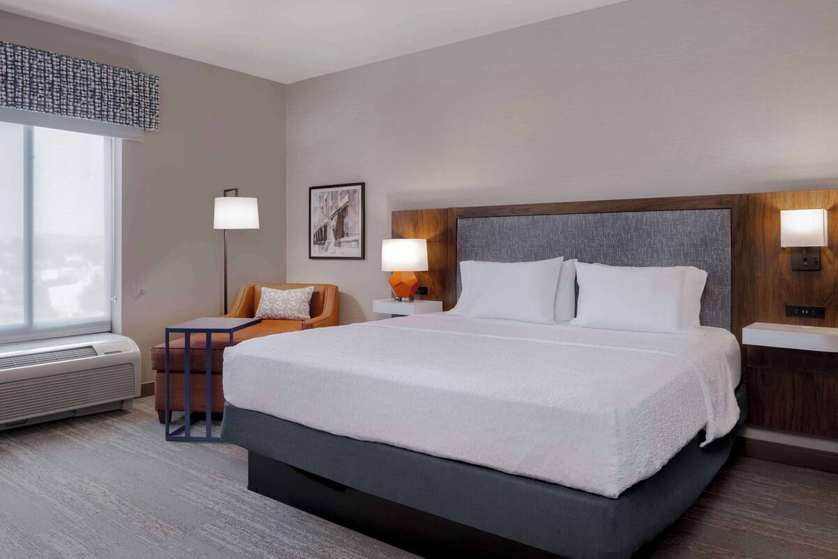A hotel room with a large bed, a desk, a chair, two wall-mounted lamps, and a large window.