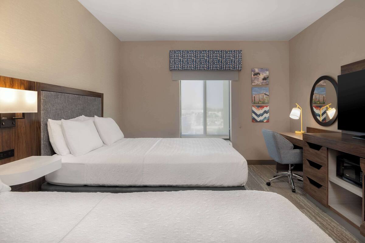 A hotel room with two beds, a desk, a chair, a TV, and artwork on the wall.