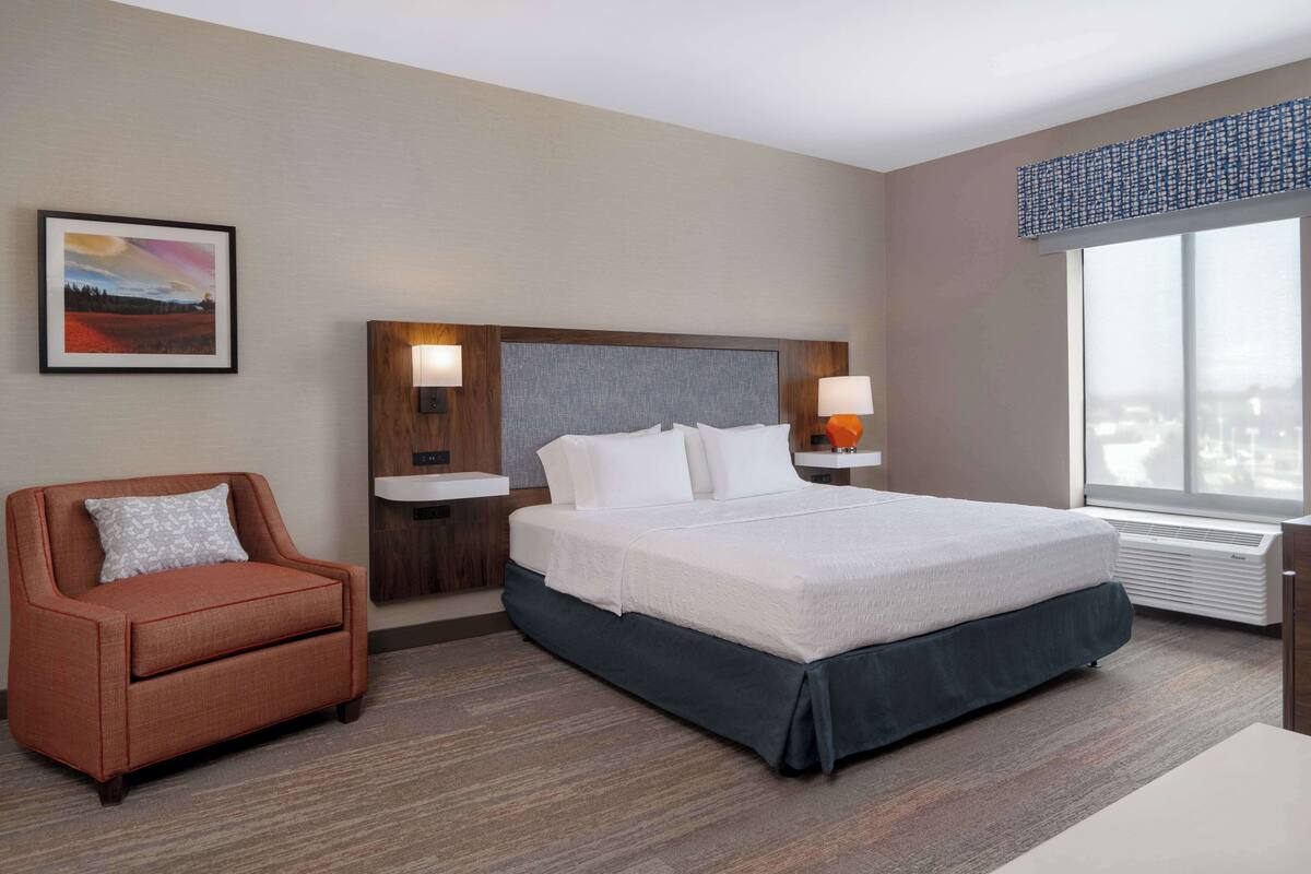 A hotel room with a bed, a chair, a nightstand, a lamp, a window, and a painting on the wall.