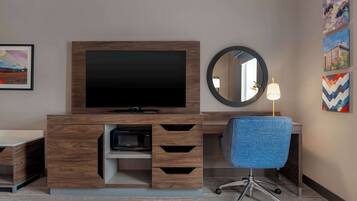 Une chambre d’hôtel moderne avec un meuble de rangement en bois, un téléviseur à écran plat, un miroir rond, une chaise de bureau bleue et des œuvres d’art accrochées aux murs.