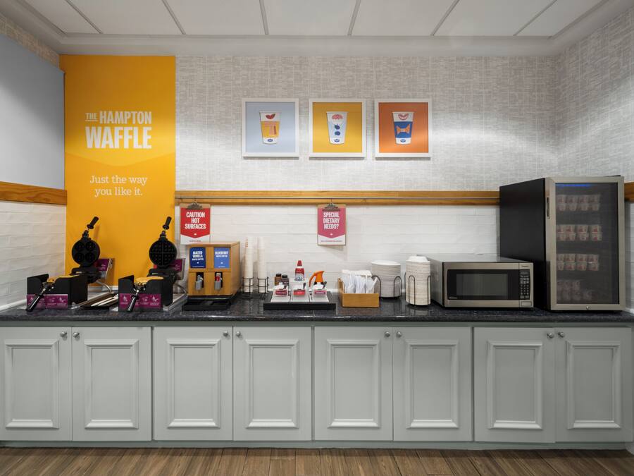A kitchenette with a waffle maker, microwave, and coffee maker. There are signs for dietary needs and caution about hot surfaces.