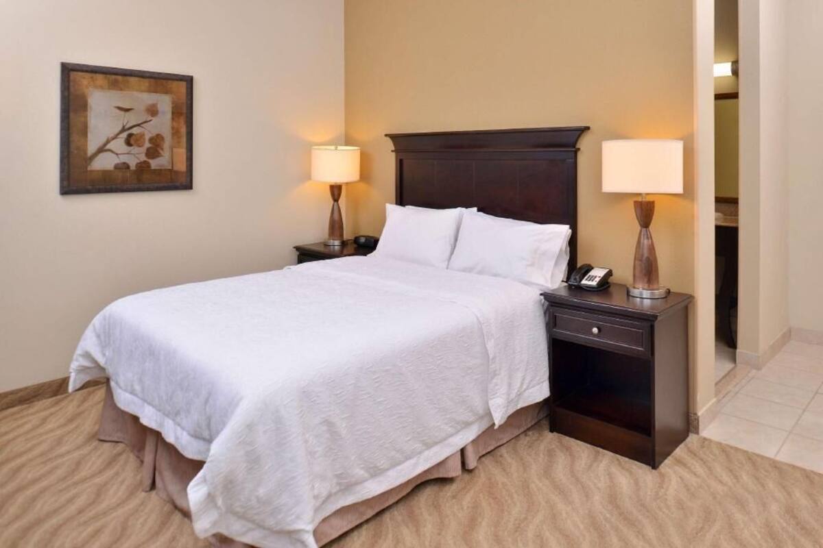 Une chambre d’hôtel avec un lit, deux tables de chevet, une petite table, une lampe et un tableau encadré au mur.