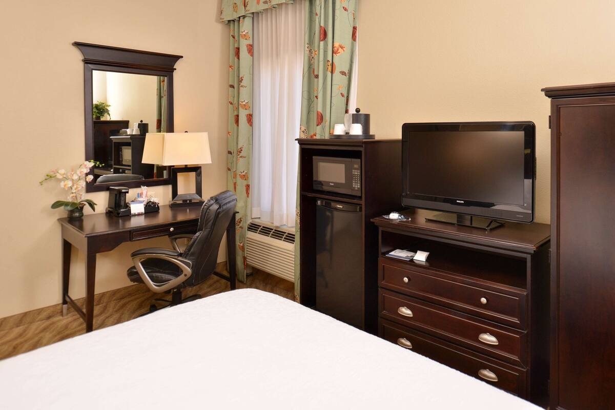 Une chambre d’hôtel comprenant un lit, un bureau avec une chaise, une télévision, une commode, un miroir et une fenêtre avec des rideaux.