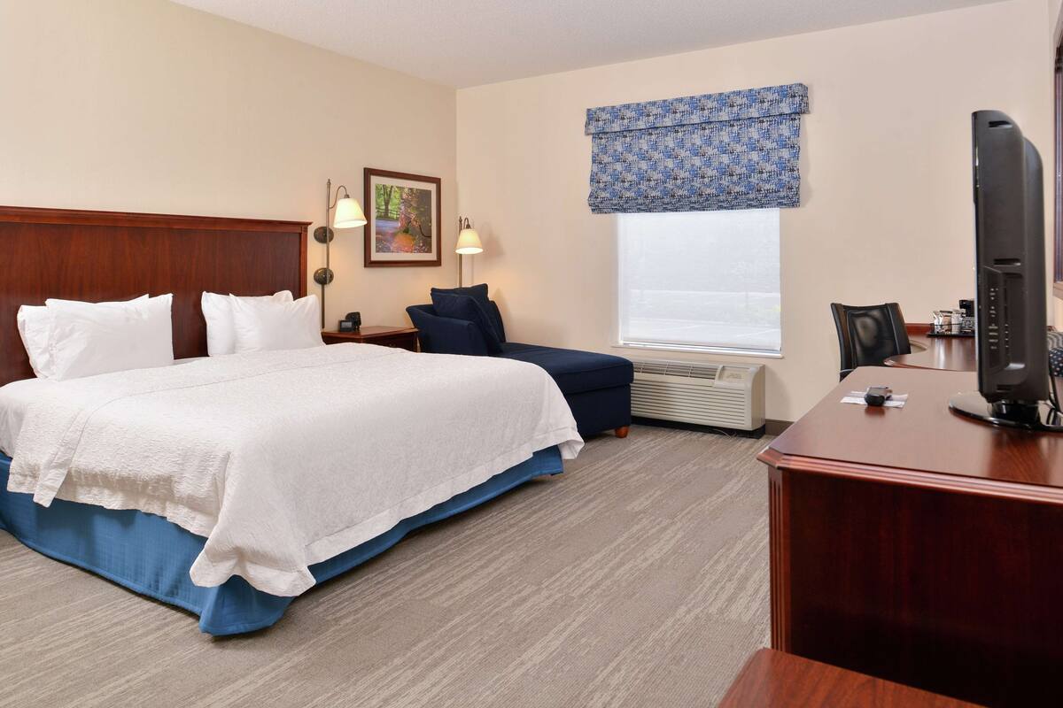 A hotel room with a large bed, a desk with a computer, a chair, a window with a blue valance, and an air conditioning unit.