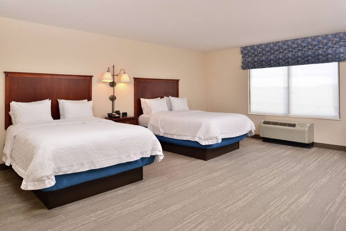 A hotel room with two beds, a nightstand with a lamp, a window with a blue valance, and a wall-mounted air conditioner.