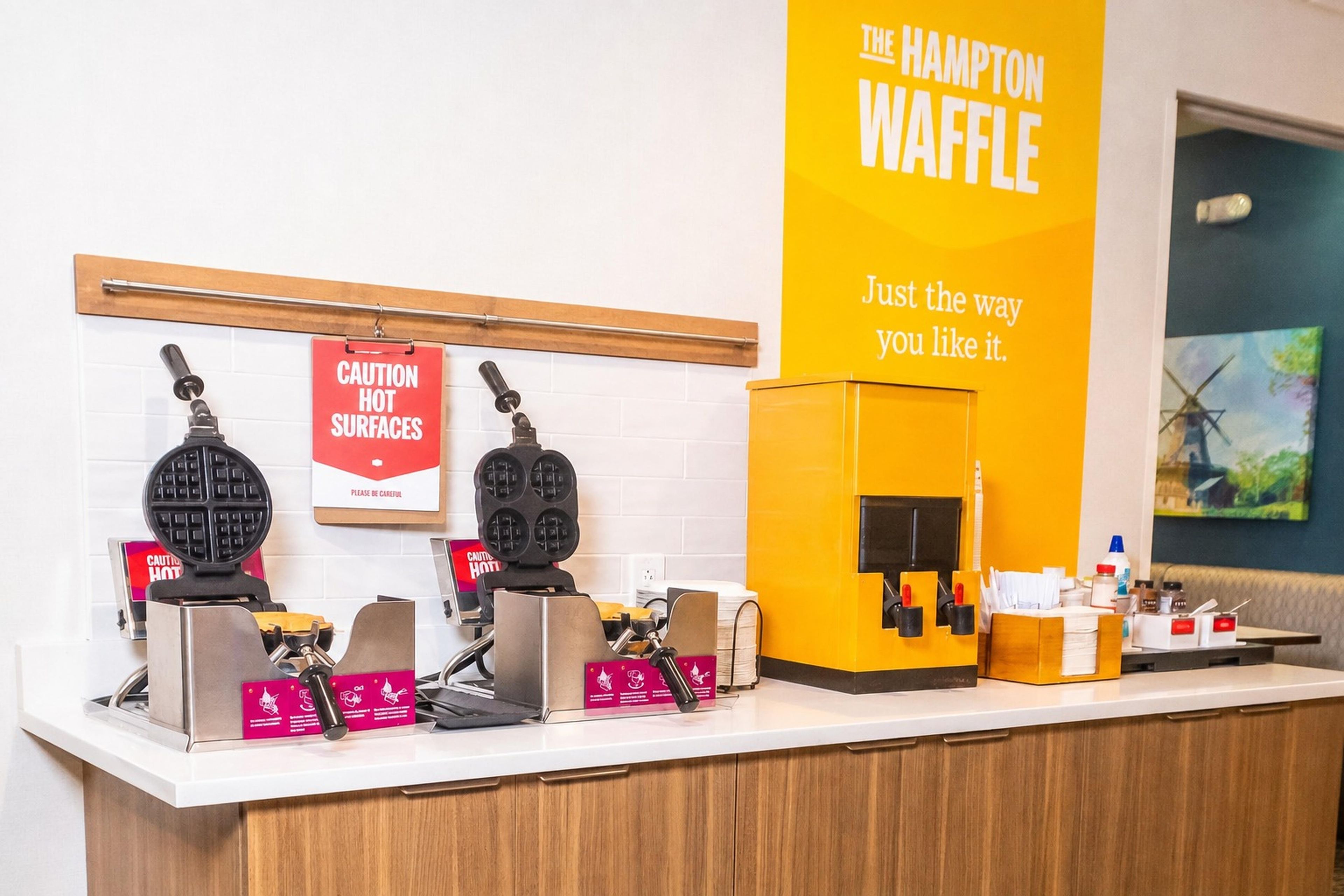 A waffle station with caution signs for hot surfaces, a sign for The Hampton Waffle, and a coffee machine.