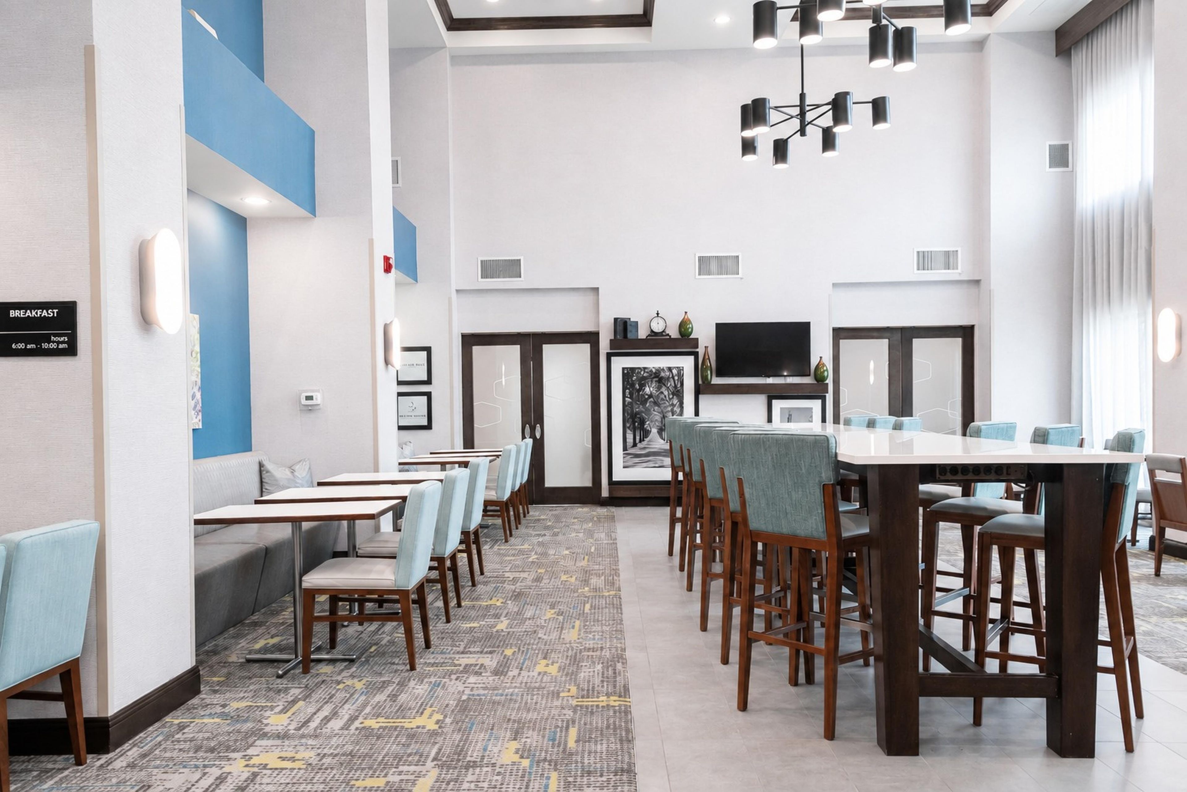 A hotel dining area with multiple tables and chairs, a television, and a sign indicating breakfast hours.