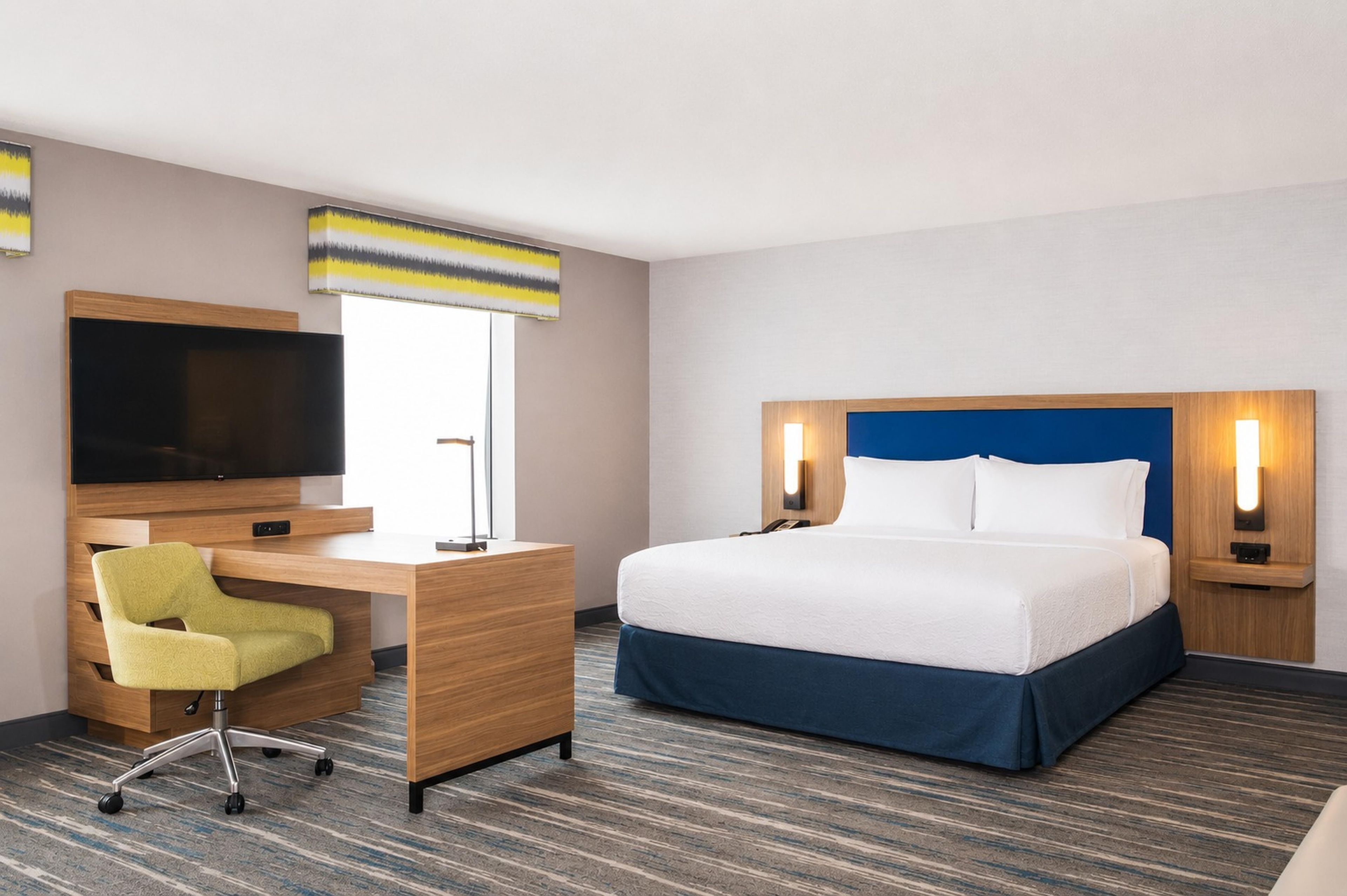 A hotel room with a bed, a desk, a chair, a TV, and a window with a striped curtain.