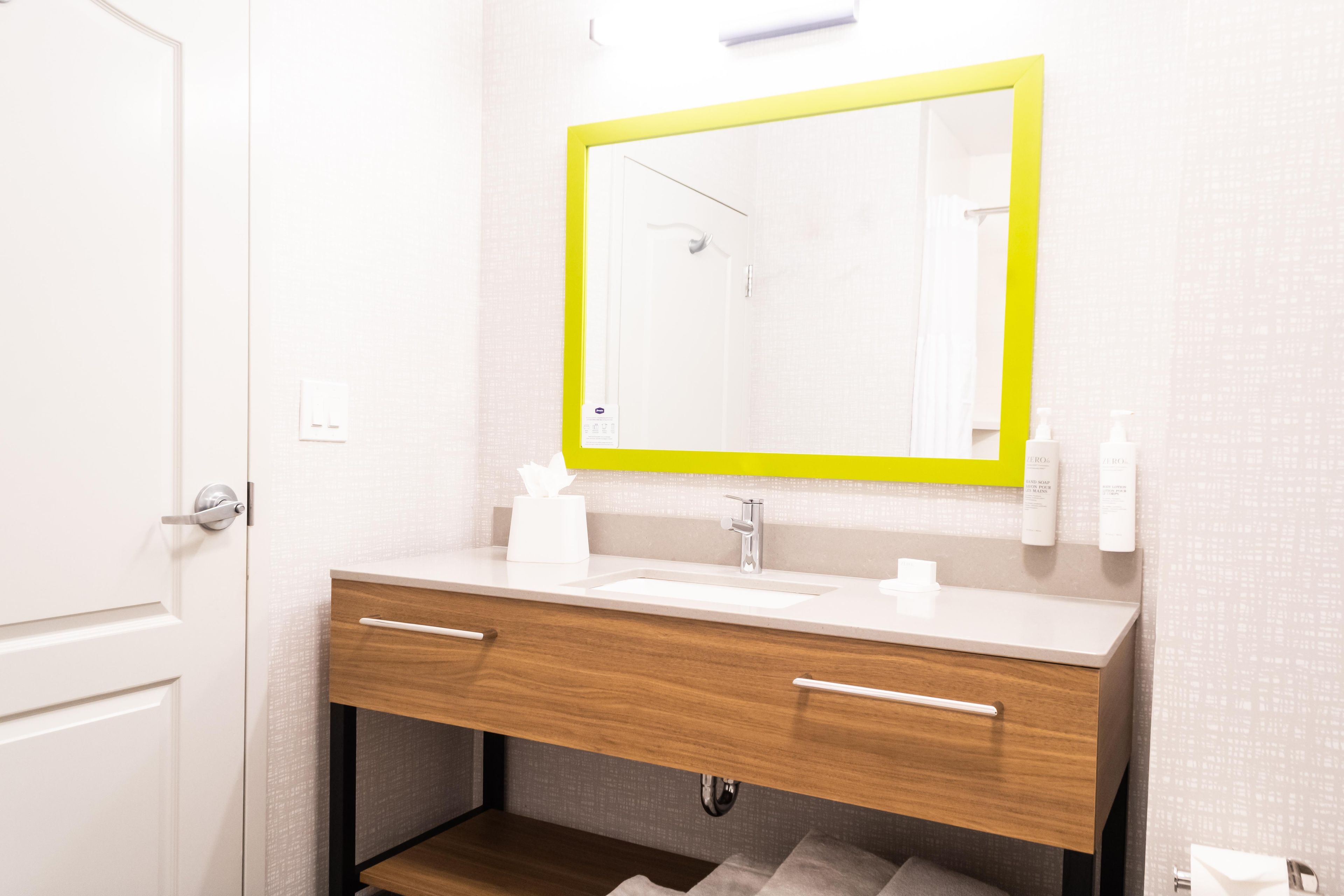 Un baño con un lavamanos blanco, tocador de madera, un espejo con marco amarillo y artículos de aseo en la pared.