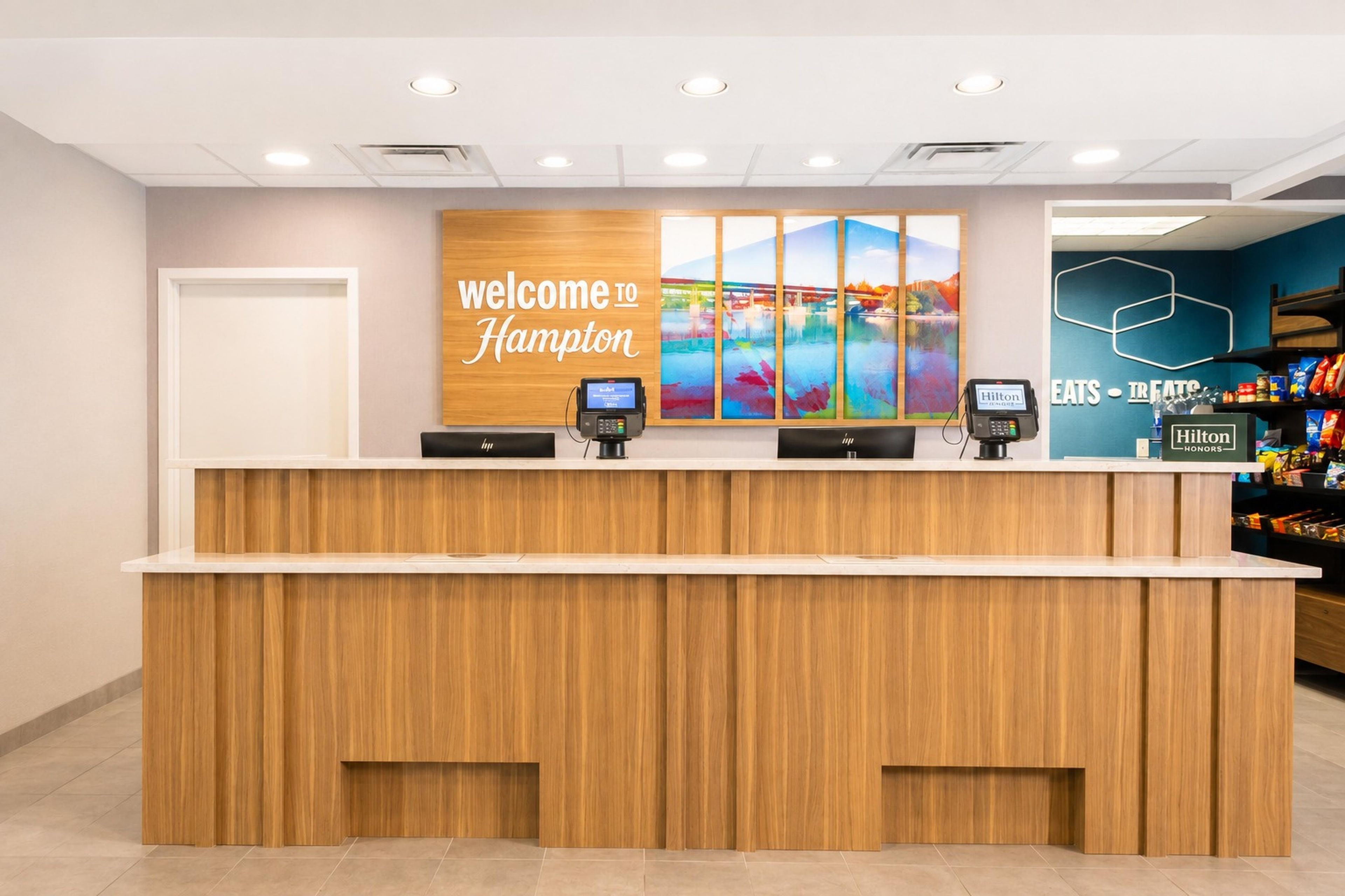 A hotel reception desk with a 'Welcome to Hampton' sign, two digital kiosks, and a wall with a scenic artwork.