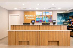 Una recepción de hotel con un cartel que dice 'Welcome to Hampton', dos quioscos digitales y una pared con una obra de arte de paisaje.