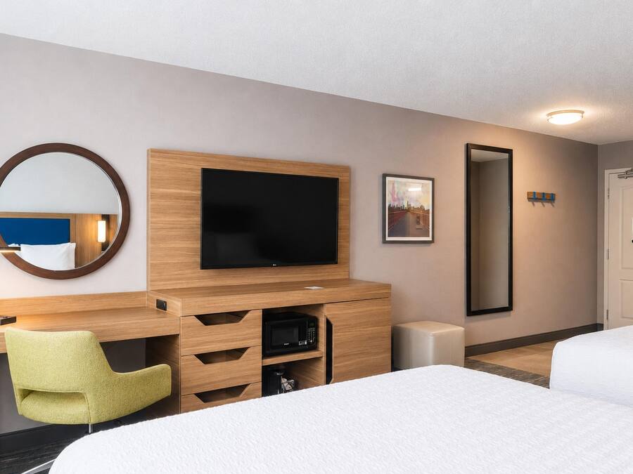 A hotel room with a bed, a desk with a television, a mirror, a chair, and a door.