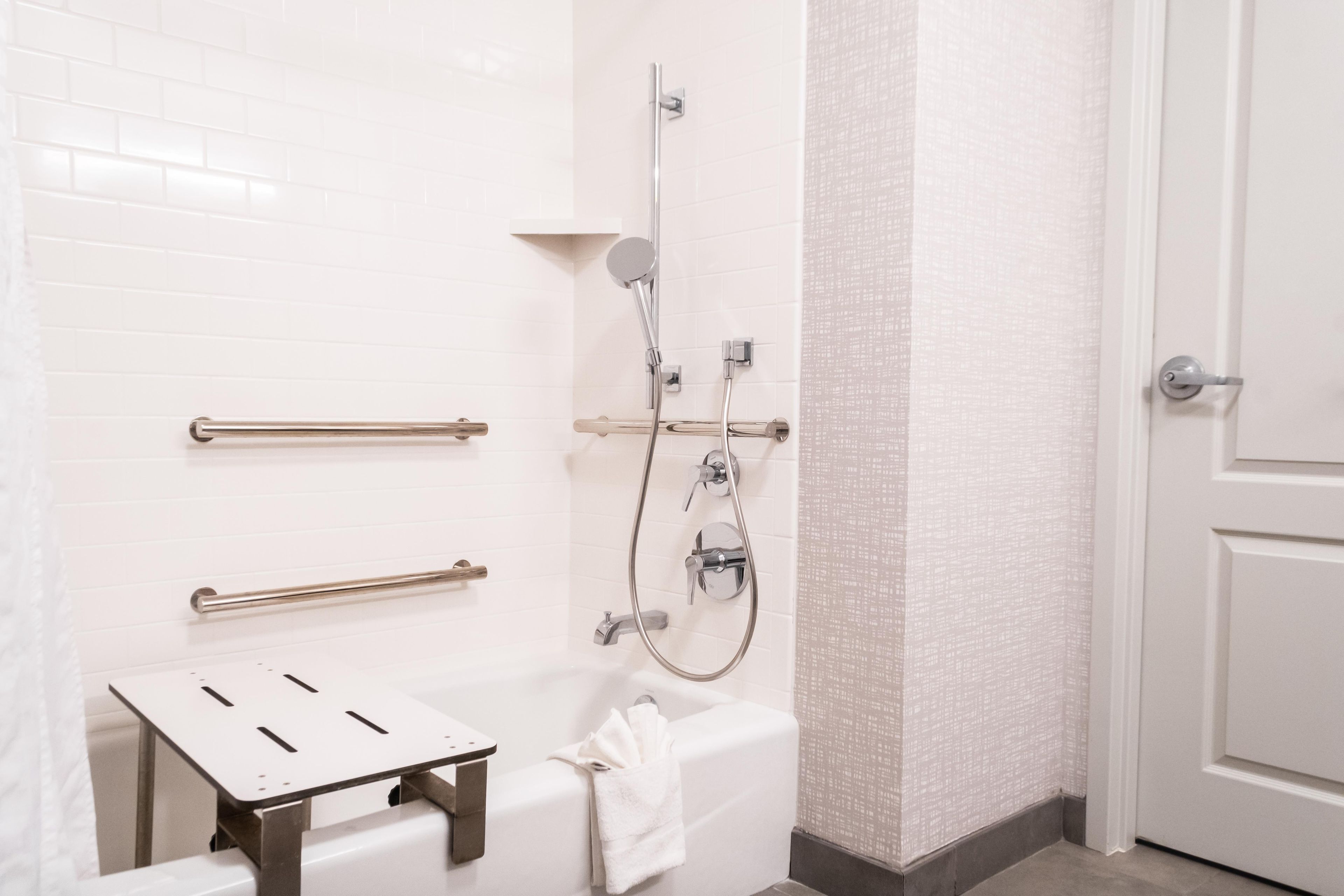 A bathroom with a white tiled wall, a shower with a handheld showerhead, a grab bar, a towel rack, and a toilet.