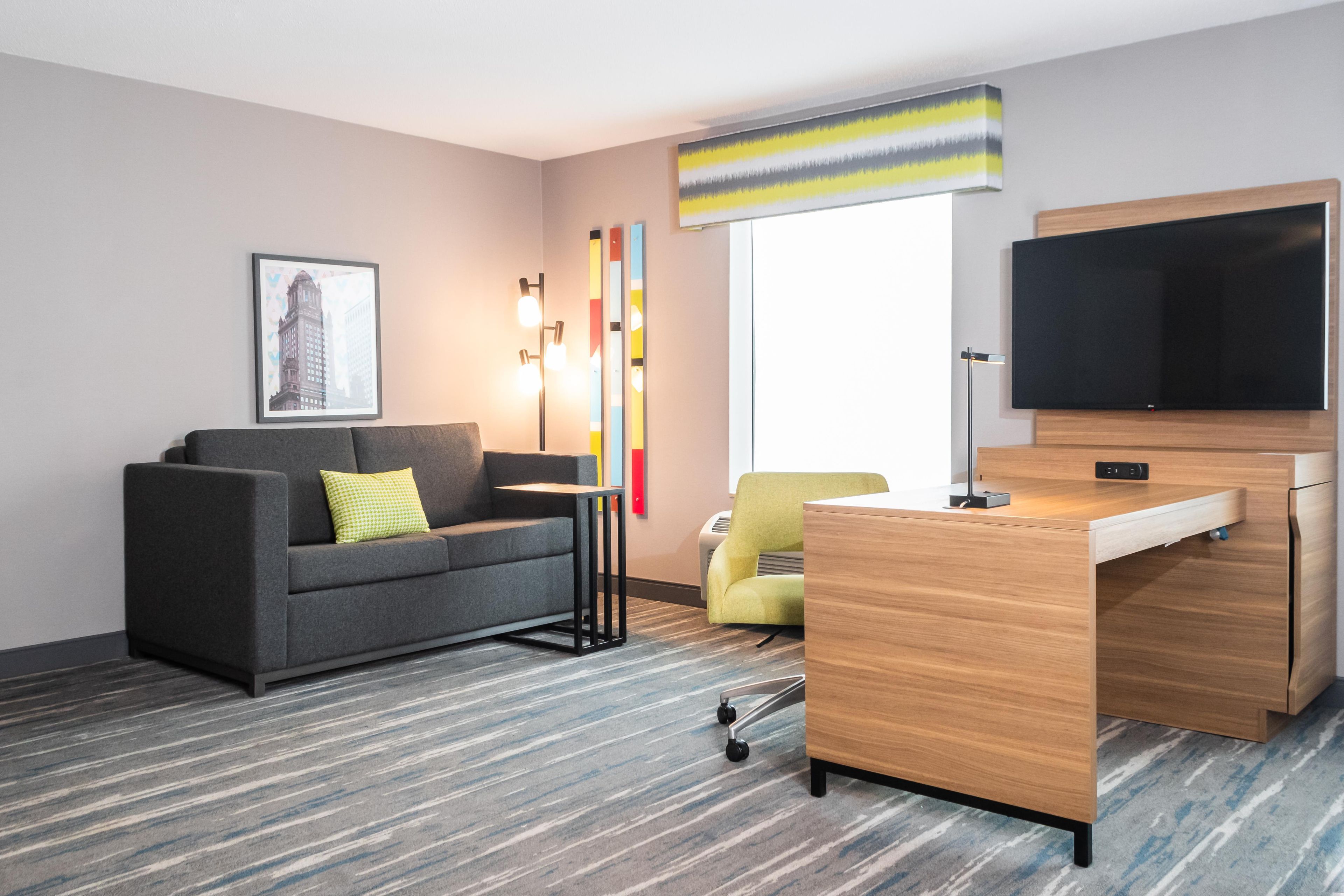 A modern hotel room with a gray sofa, a small desk, a flat-screen TV, and a window with a striped curtain.