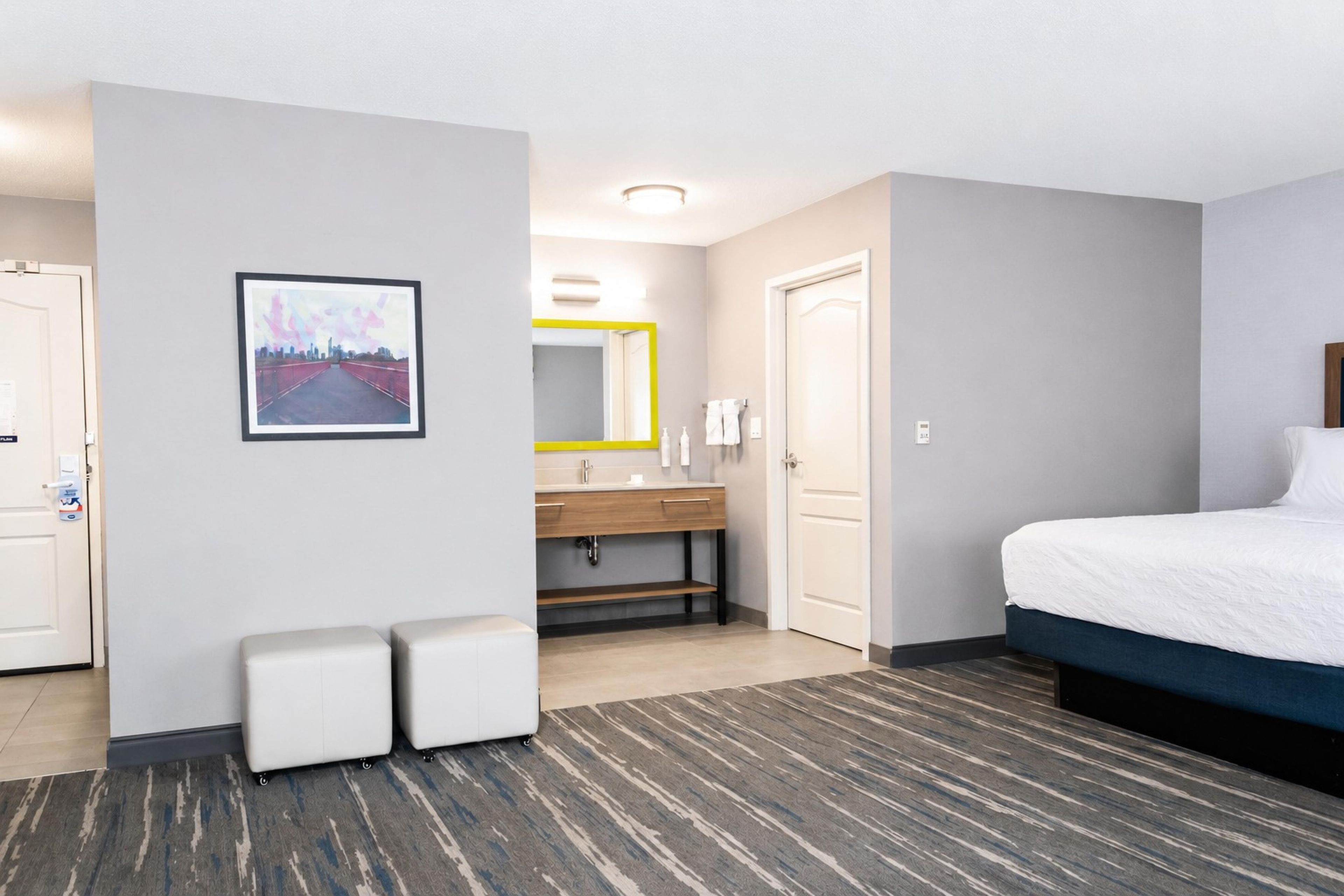 A hotel room with a bed, a bathroom with a mirror, a sink, and a toilet. There is a framed picture on the wall and a carpeted floor.
