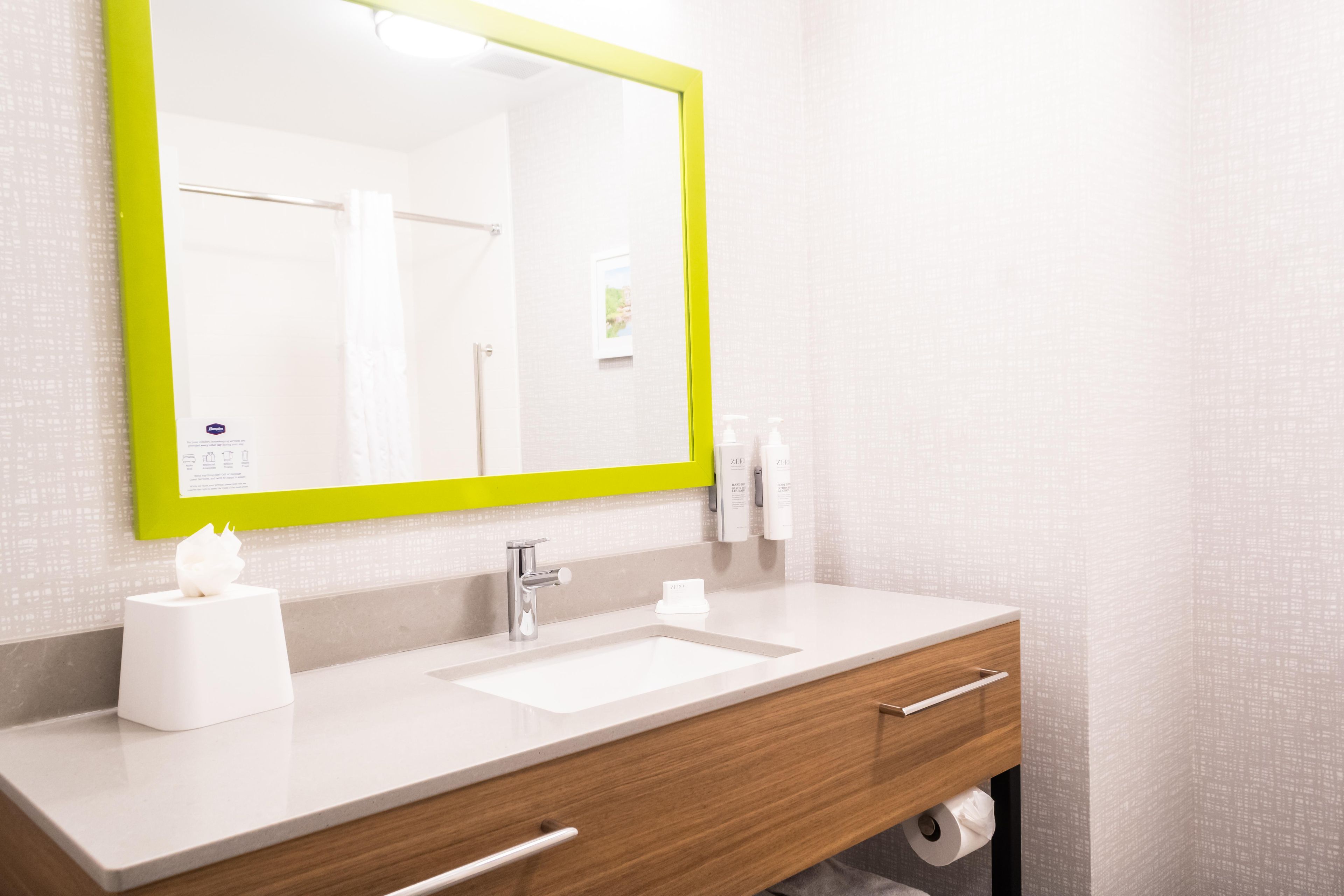 A bathroom with a white sink, a mirror with a green frame, a towel dispenser, and a toilet paper holder.