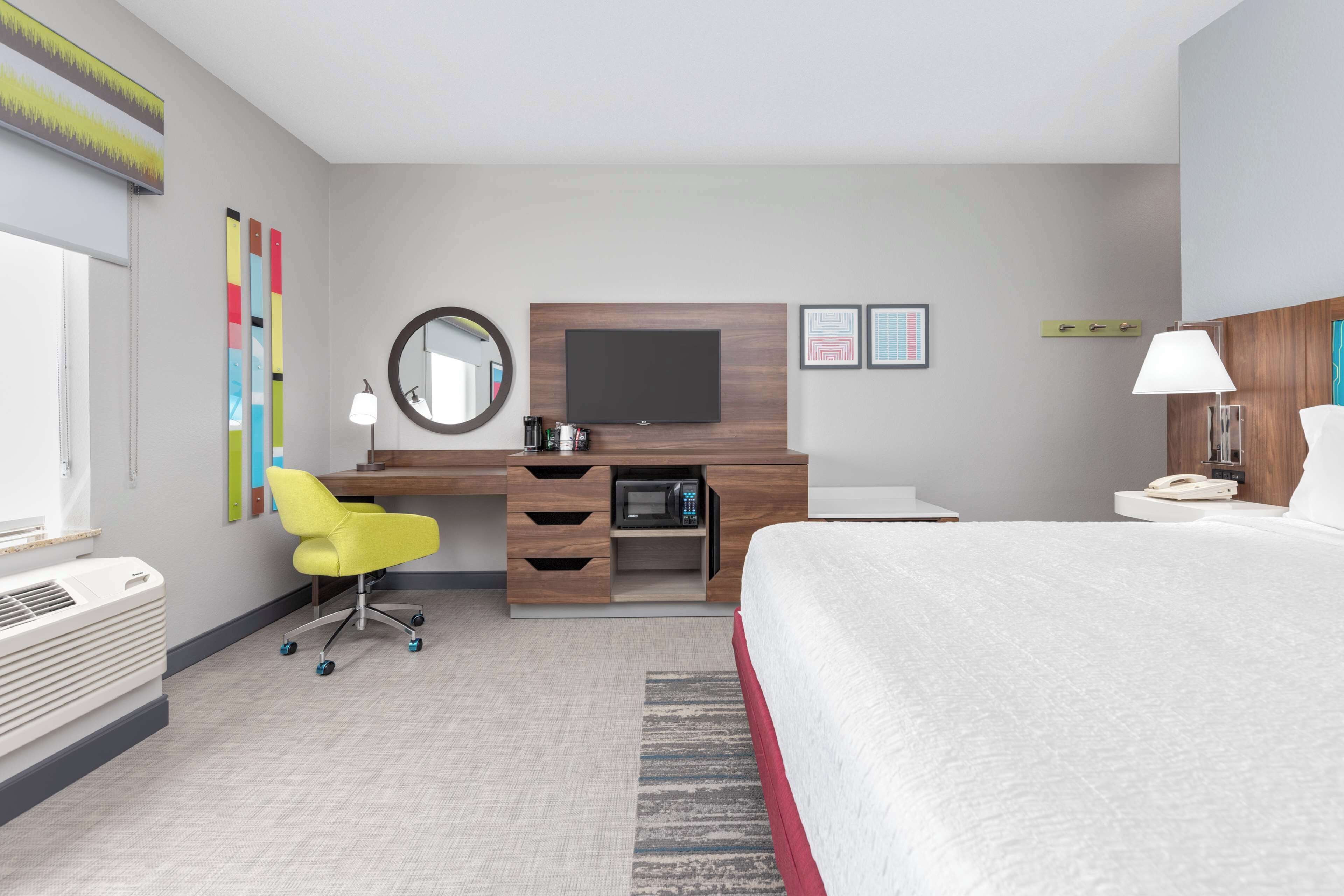 A modern hotel room with a bed, a desk with a flat-screen TV, a microwave, a chair, and a bedside table with a lamp.