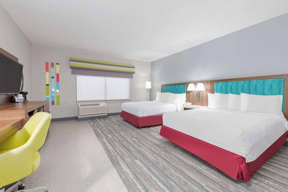 A hotel room with two beds, a desk, a chair, a TV, and a window.