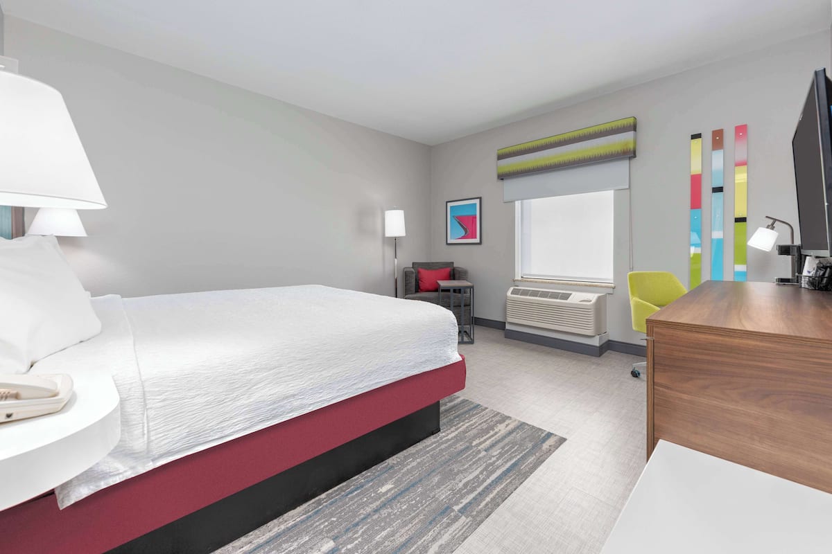 A modern hotel room with a large bed, a desk, a chair, a lamp, a TV, and a window with blinds.