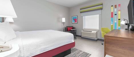 A modern hotel room with a large bed, a desk, a chair, a lamp, a TV, and a window with blinds.