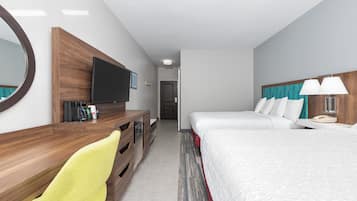 A hotel room with a wooden desk, a flat-screen TV, a bed with white bedding, a bedside lamp, and a mirror.