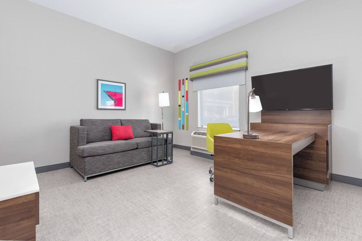 A modern hotel room with a grey sofa, a wooden desk, a flat-screen TV, a window with blinds, and a colorful abstract painting.
